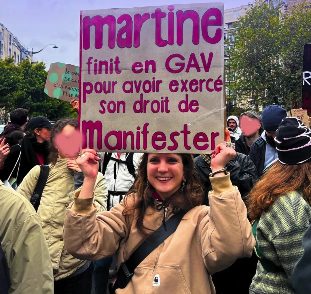 Une manifestante souriante brandit une pancarte en carton « Martine finit en GAV pour avoir exercé son droit de manifester », en lettres violettes