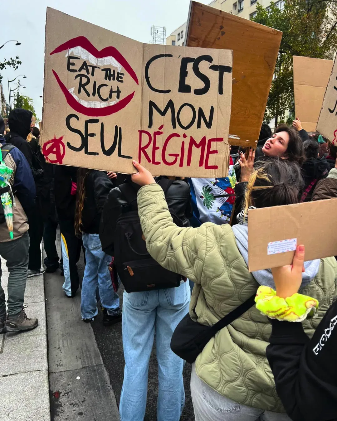 Une jeune femme en doudoune brandit deux pancartes : une bouche rouge « Eat the rich » et « C’est mon seul régime »