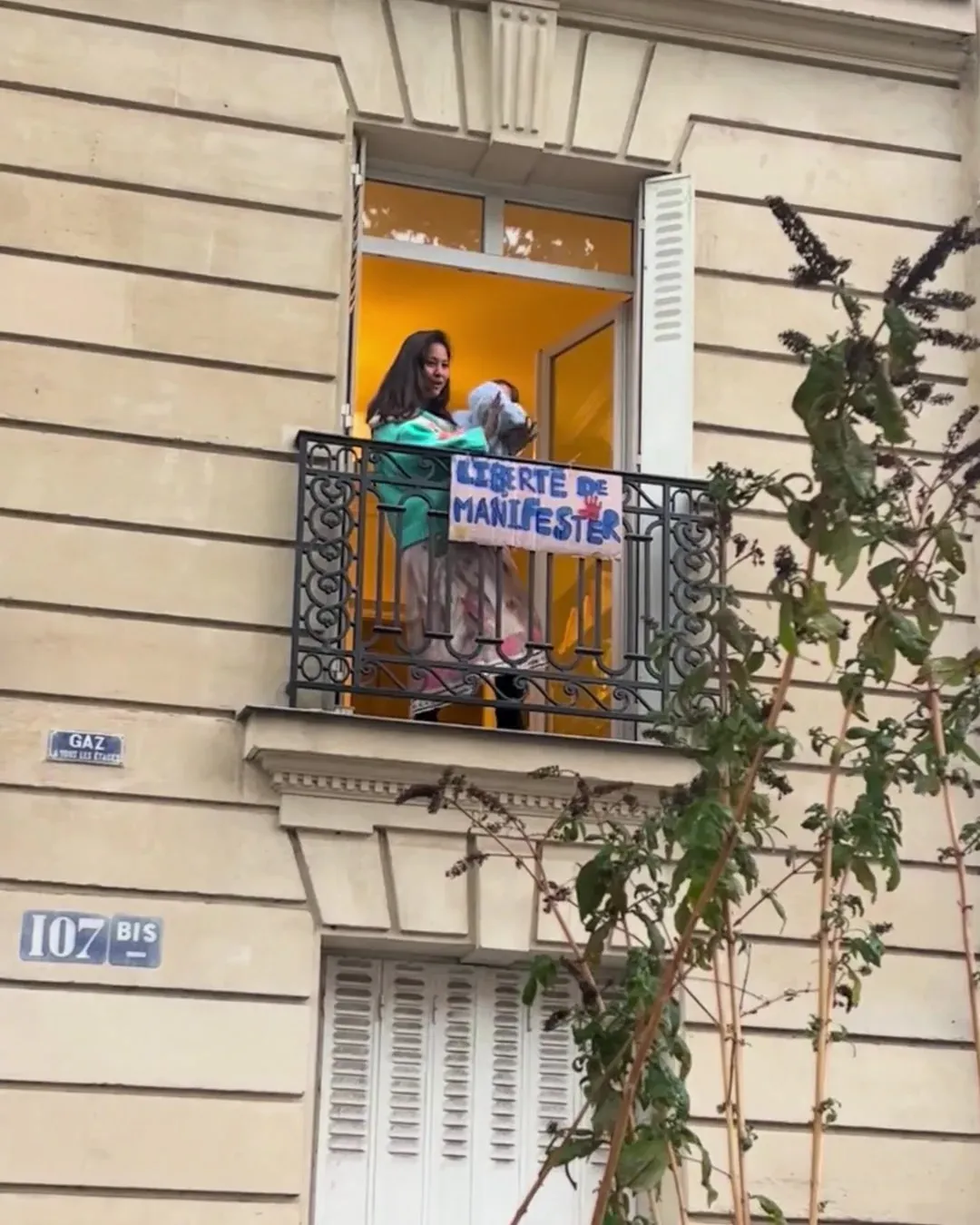 Une femme sur un balcon parisien tient un bébé dans les bras, une pancarte « Liberté de manifester » accrochée à la rambarde