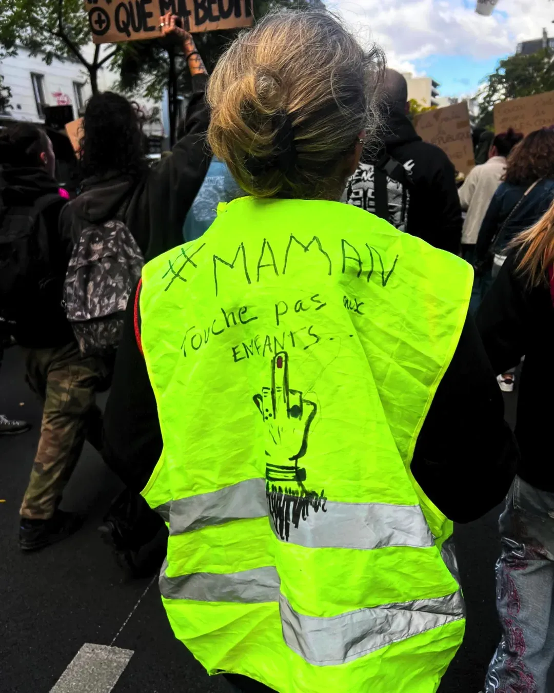 Une femme de dos porte un gilet jaune sur lequel est écrit au feutre « #Maman, touche pas aux enfants » avec un doigt d’honneur dessiné