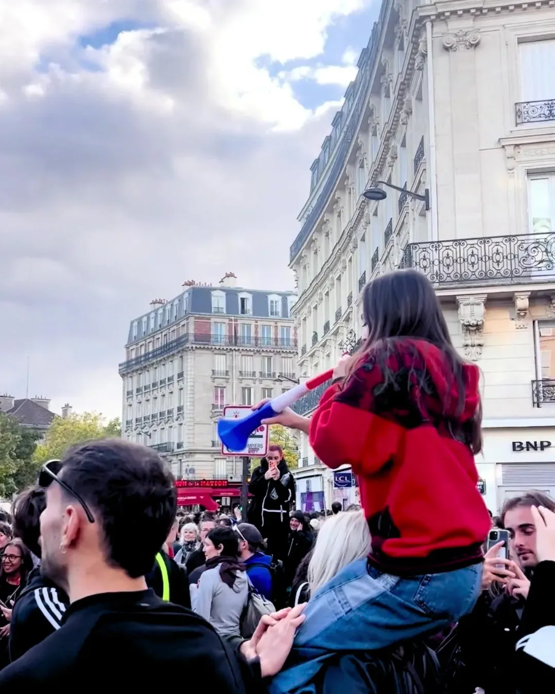Une jeune femme en sweat rouge sur les épaules d’un ami, souffle dans une vuvuzela bleu, blanc, rouge, au milieu de la foule