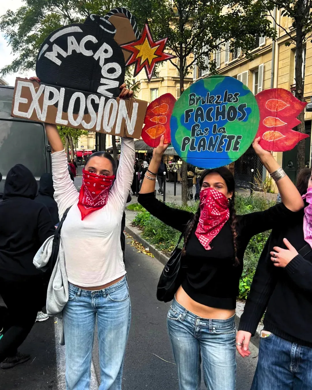 Deux jeunes femmes avec des bandanas rouges brandissent une pancarte « Macron Explosion » avec une bombe dessinée et une planète « Brûlez les fachos, pas la planète »