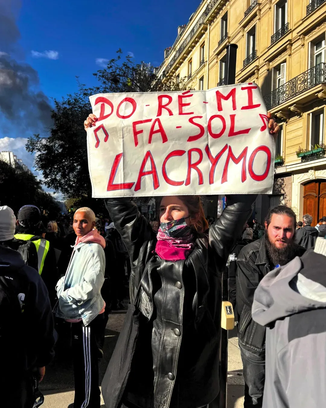Une manifestante en veste de cuir brandit une pancarte « Do ré mi fa sol lacrymo » avec des notes de musique, sous un ciel bleu