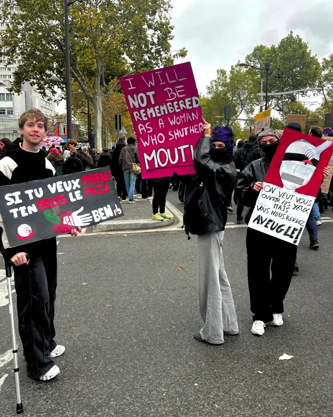 Trois amies côte à côte sur la chaussée avec leurs pancartes : « I will not be remembered as a woman who shut her mouth », une pancarte sur les lanceurs de balles de défense et une troisième « On veut vous ouvrir les yeux mais vous nous rendez aveugles »