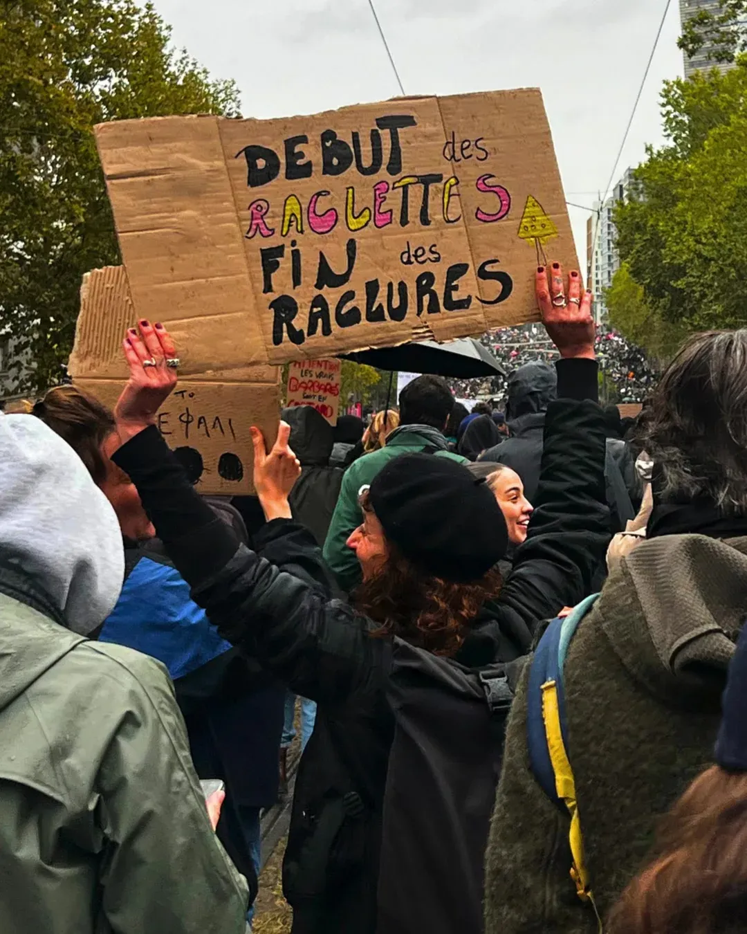 Une manifestante aux cheveux bouclés, bonnet noir, lève une pancarte en carton « Début des raclettes, fin des raclures »