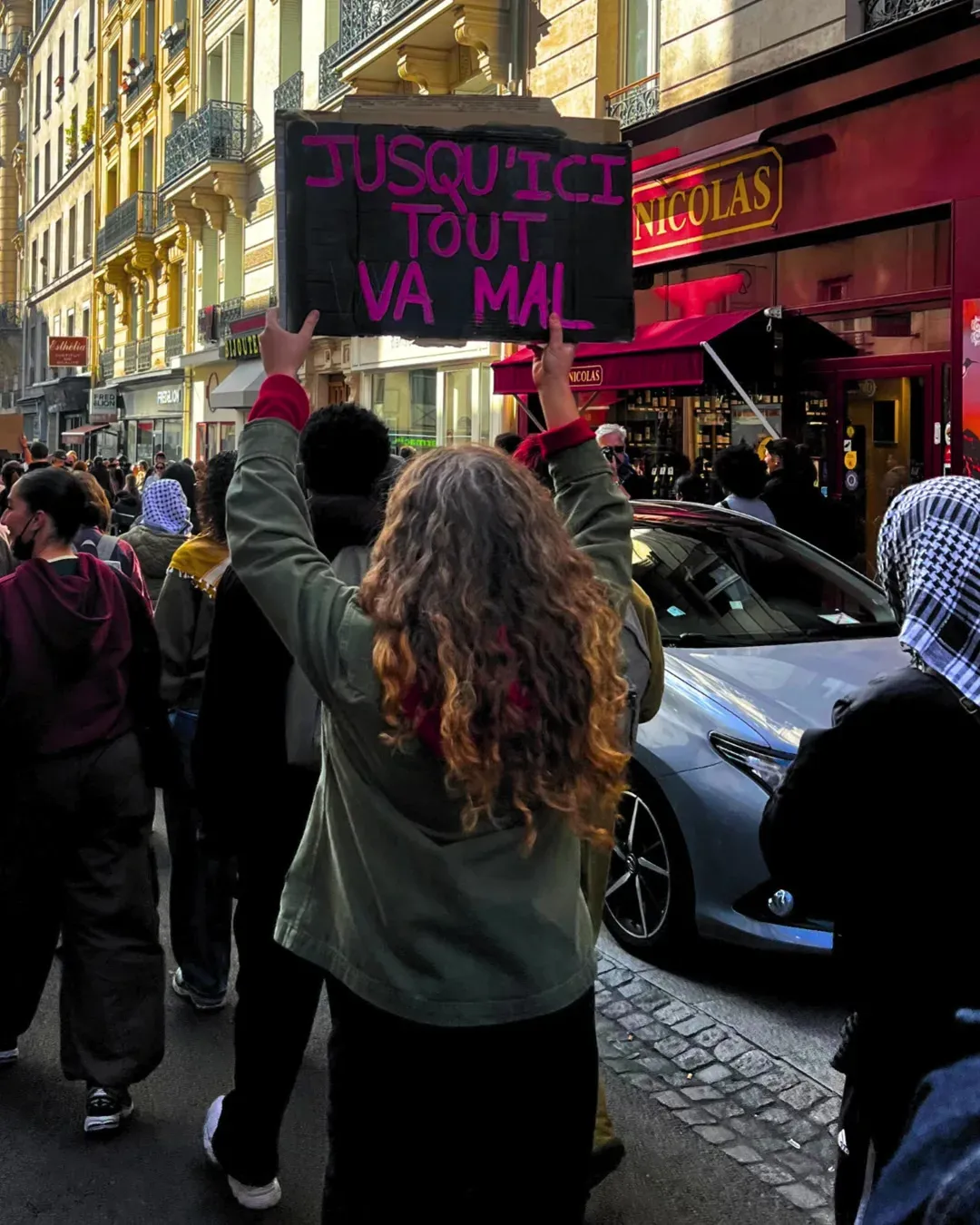 Une manifestante aux longs cheveux bouclés, de dos, brandit une pancarte noire « Jusqu’ici tout va mal » en lettres roses dans une rue de Paris
