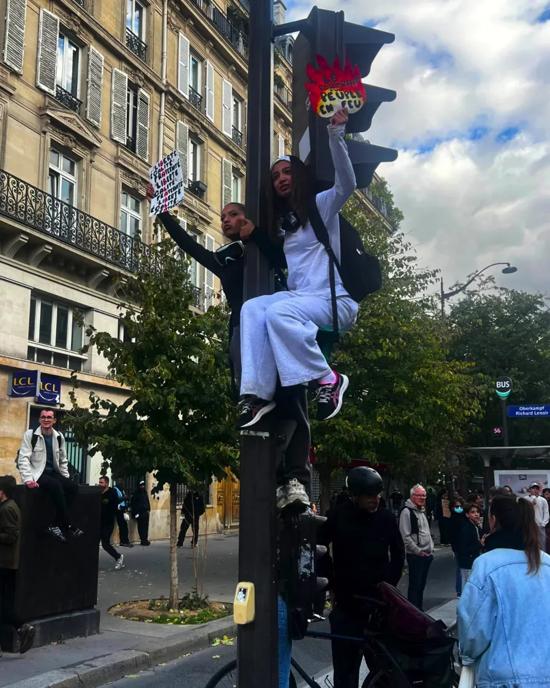 Deux jeunes femmes assises tout en haut d’un feu tricolore, pancartes à la main, au-dessus du cortège
