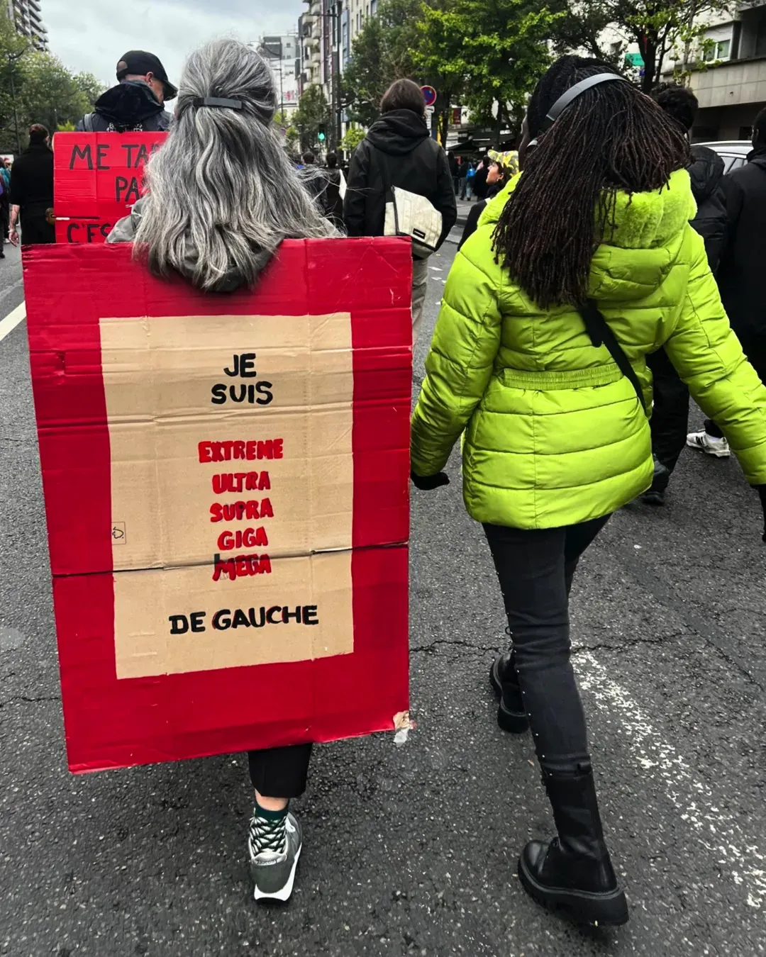 Deux femmes de dos, l’une aux cheveux gris avec une pancarte rouge « Je suis extrême ultra supra giga méga de gauche », l’autre en doudoune vert fluo