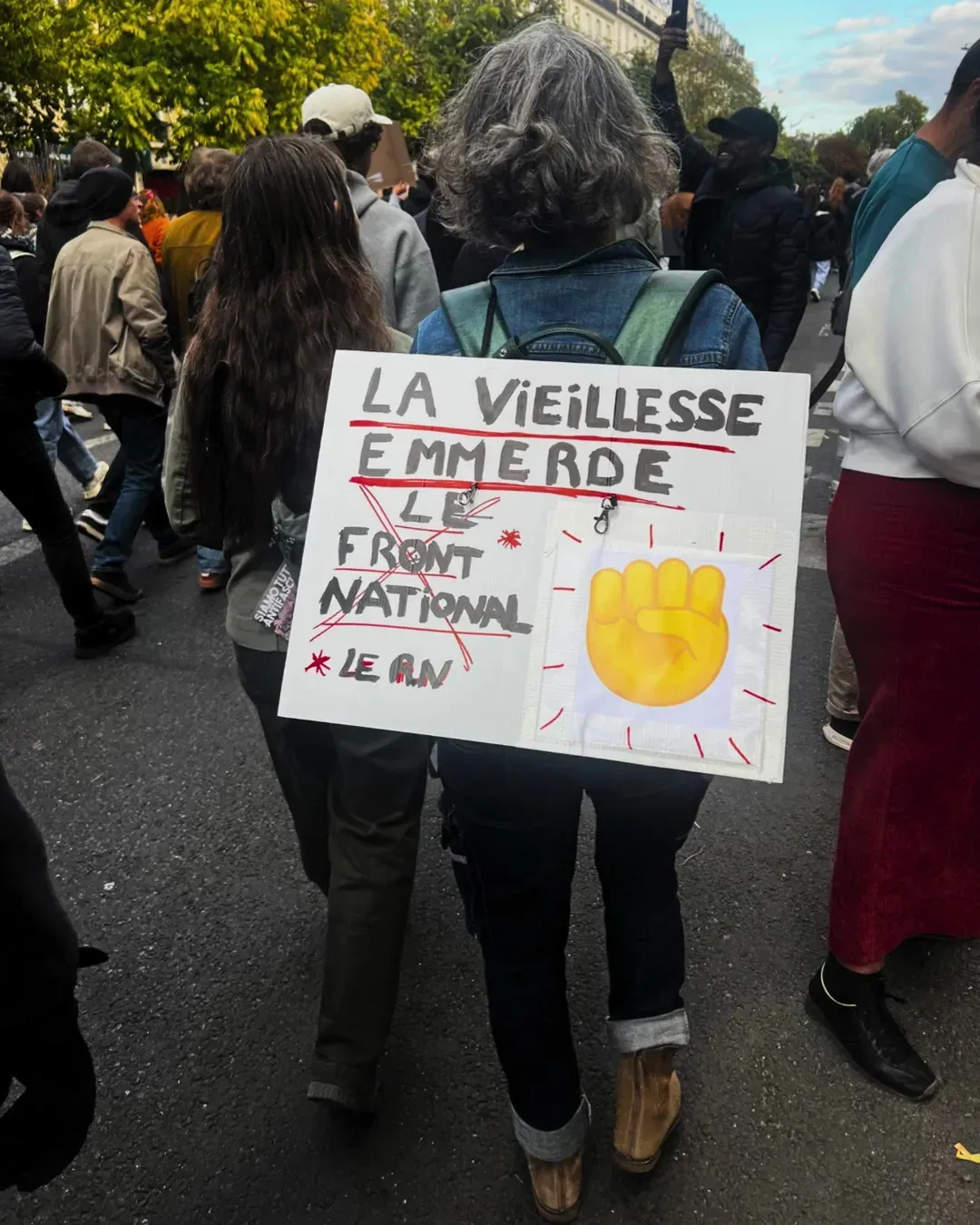 Une femme aux cheveux gris, de dos, porte sur son sac une pancarte « La vieillesse emmerde le Front National, le RN » avec un poing levé jaune