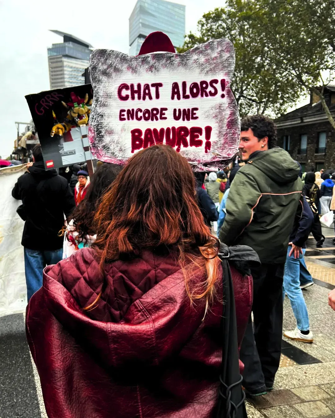 Une manifestante aux cheveux roux, de dos, tient une pancarte en forme de tête de chat « Chat alors ! Encore une bavure ! »