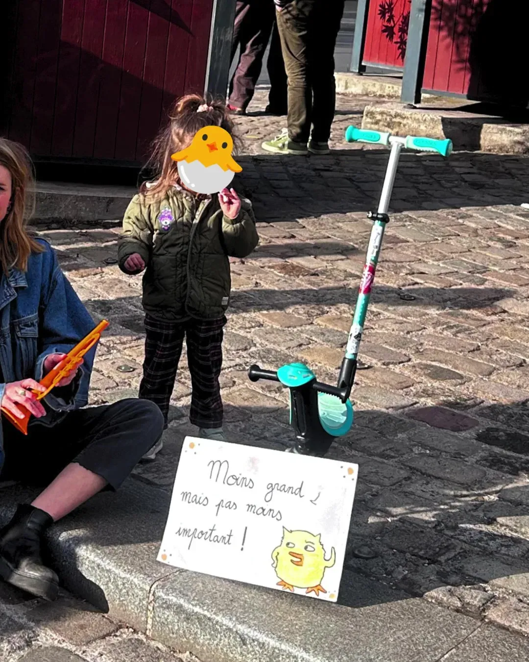Une petite fille, visage caché par un poussin, debout à côté de sa trottinette et d’une pancarte « Moins grand, mais pas moins important ! », sa mère assise à côté