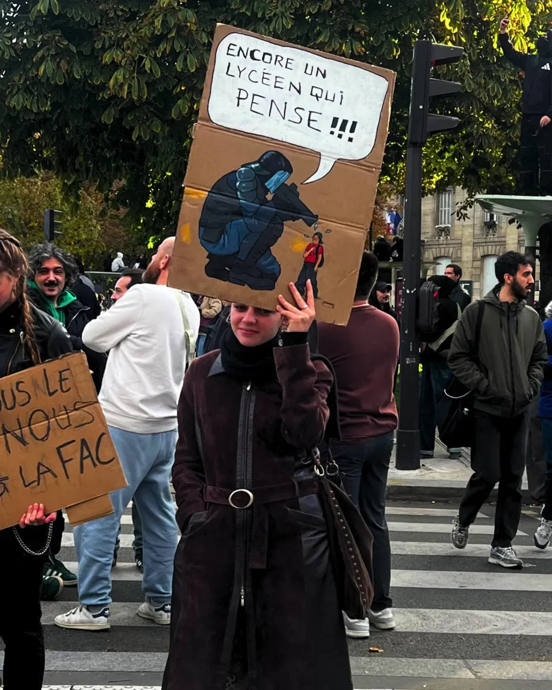 Une manifestante en manteau brun brandit une pancarte avec un policier anti-émeute dessiné en train de viser et la bulle « Encore un lycéen qui pense !!! »