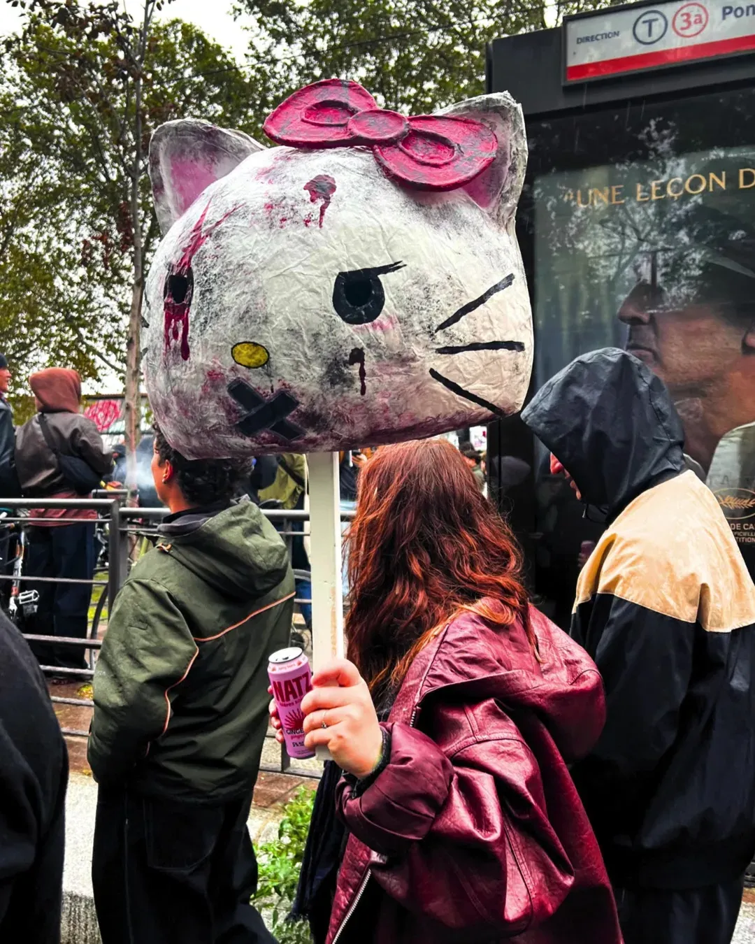 Une manifestante aux cheveux roux porte une grande tête de Hello Kitty en papier mâché, un œil crevé et ensanglanté, nœud rose sur la tête