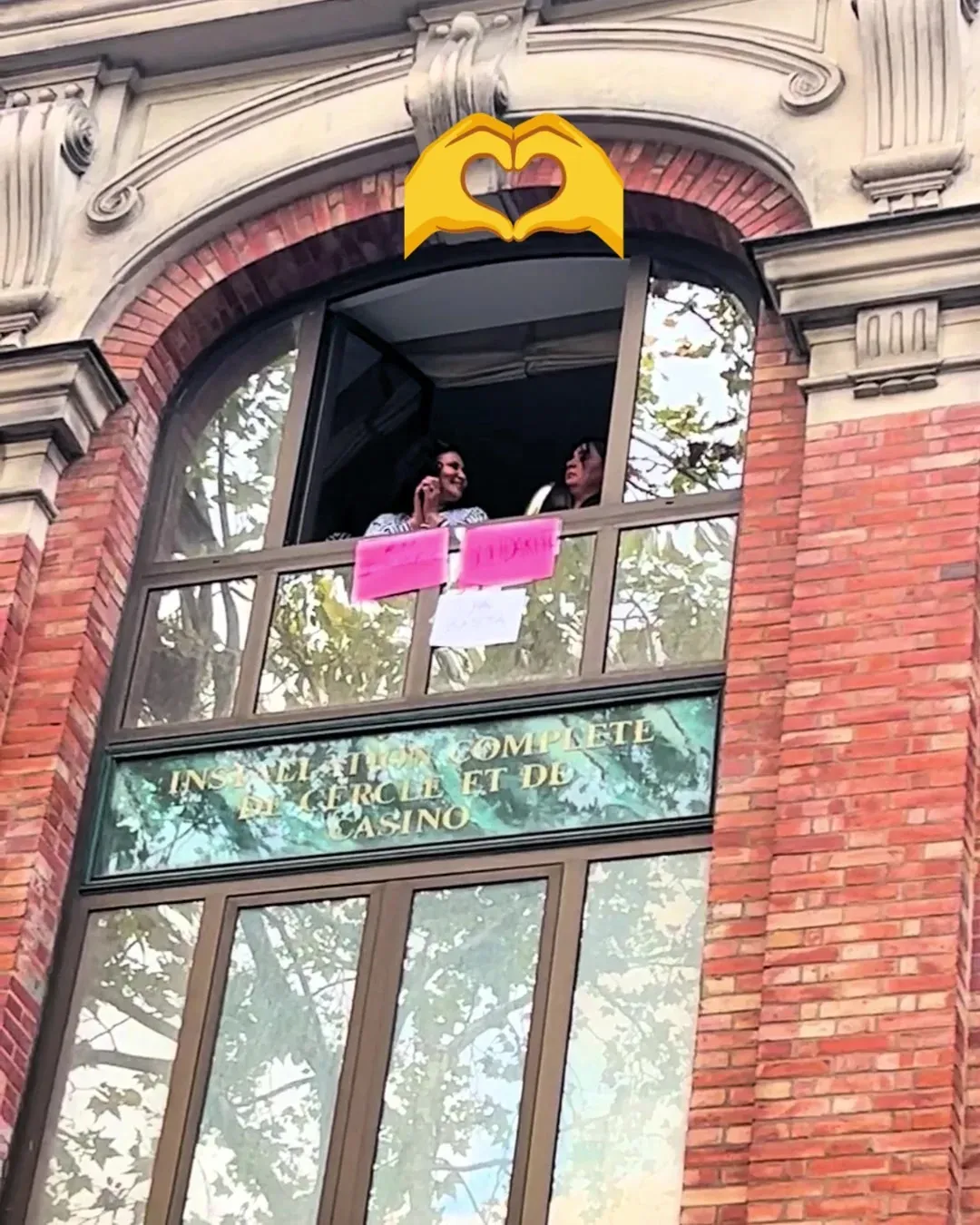 Deux femmes à la fenêtre d’un immeuble en briques rouges, des pancartes roses collées à la vitre, saluent le cortège