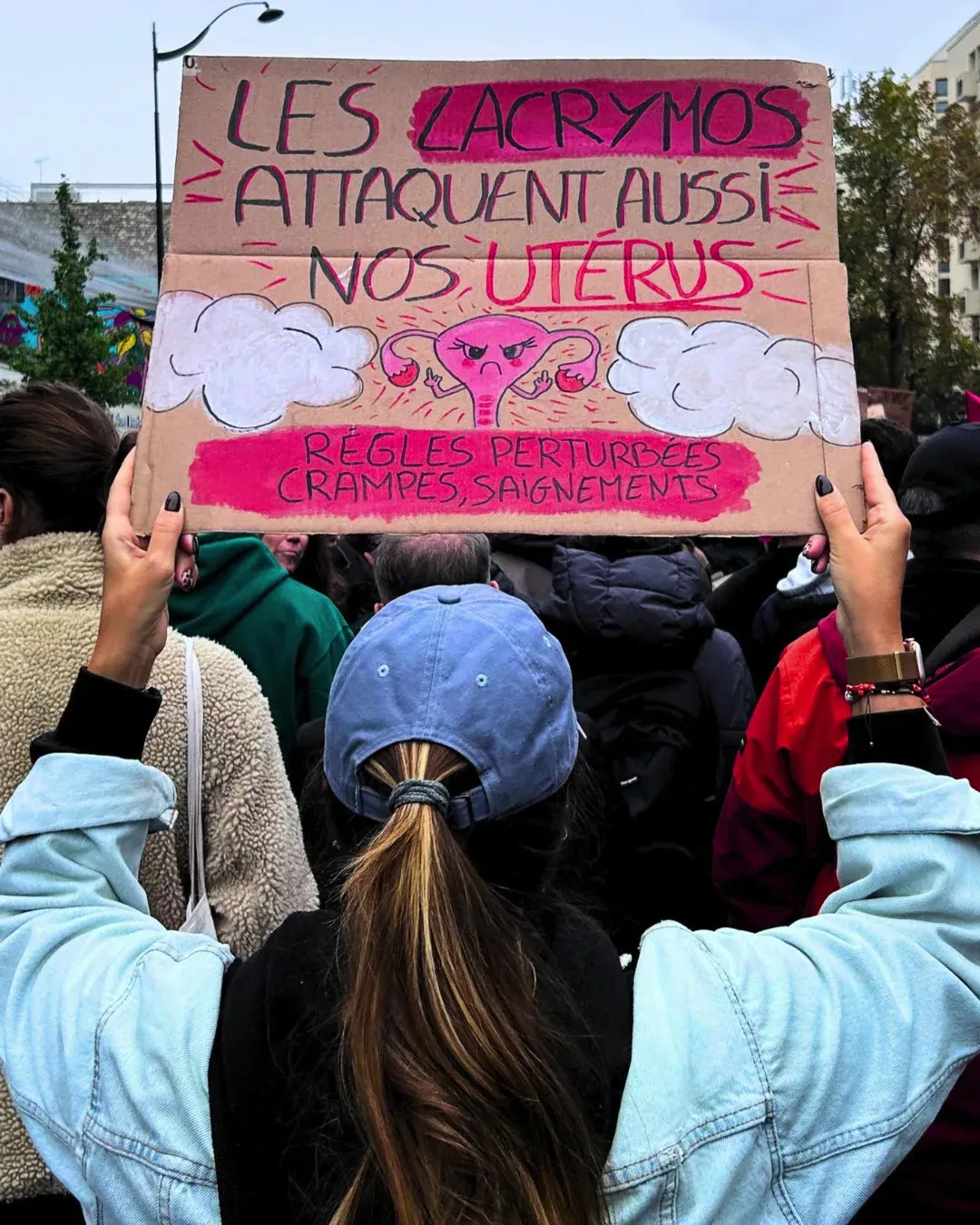 Une manifestante de dos, casquette bleue et veste en jean, lève une pancarte « Les lacrymos attaquent aussi nos utérus » avec un utérus dessiné en colère et la mention « règles perturbées, crampes, saignements »