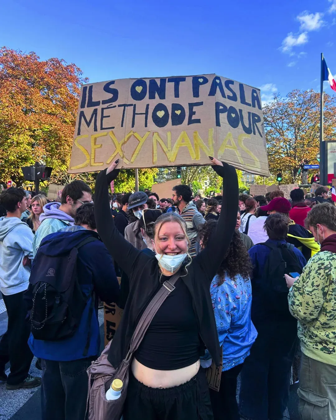 Une manifestante souriante, masque sous le menton, brandit une pancarte en carton « Ils ont pas la méthode pour sexynanas »