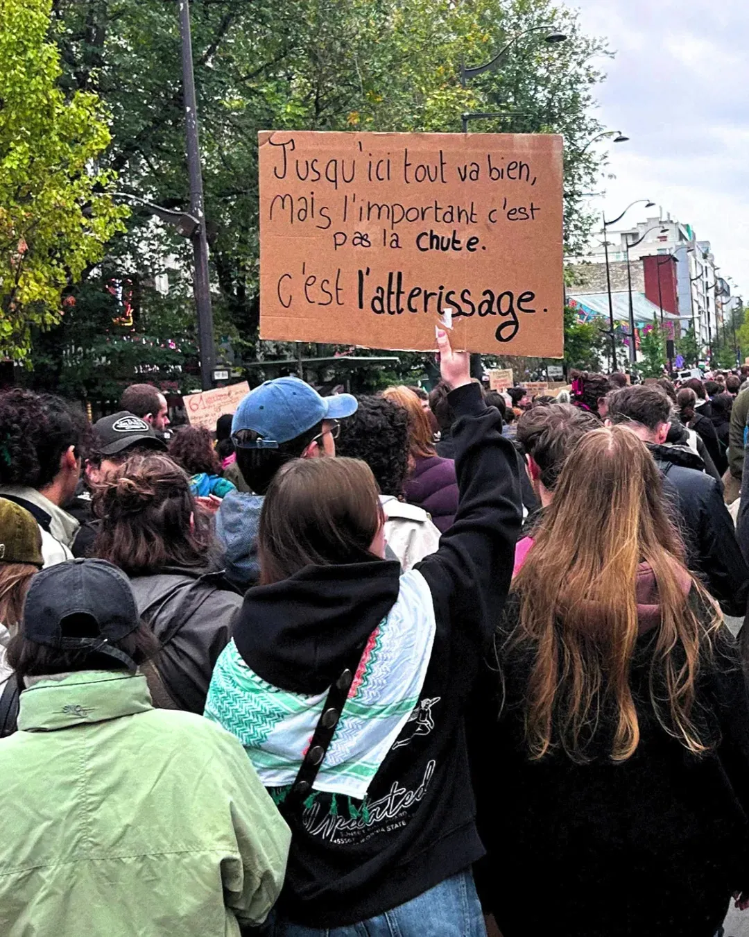 Une manifestante de dos, casquette bleue, lève une pancarte « Jusqu’ici tout va bien, mais l’important c’est pas la chute, c’est l’atterrissage »