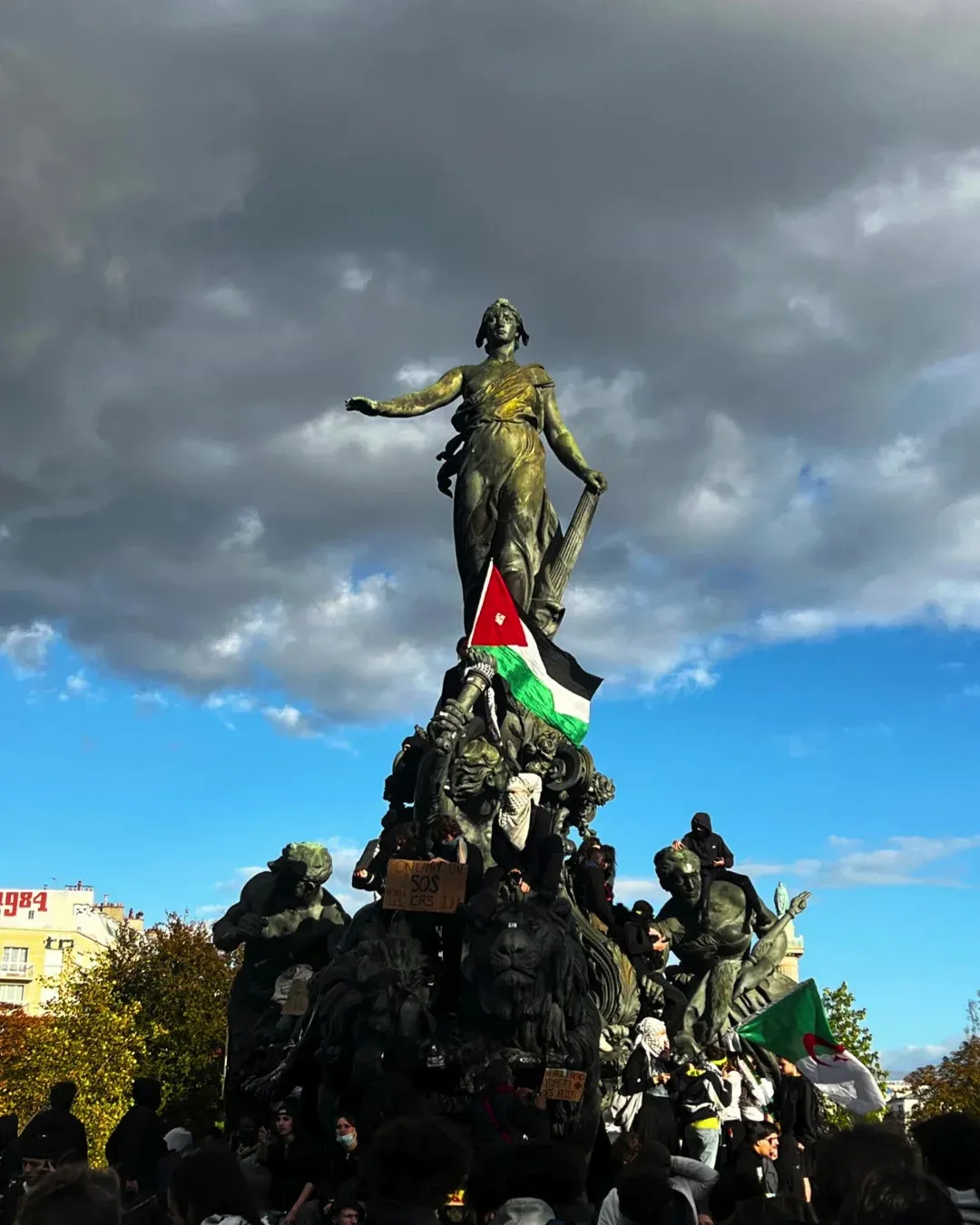 La statue du Triomphe de la République, place de la Nation, vue d’en bas, un drapeau palestinien accroché à son bras, des manifestants grimpés sur le socle sous un ciel orageux