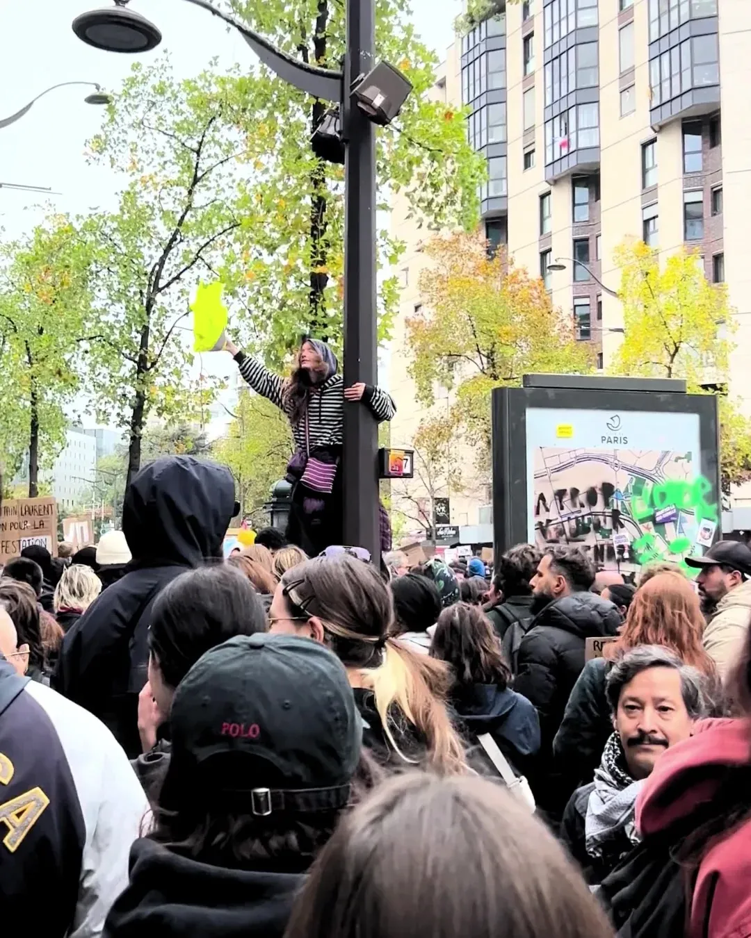 Une manifestante grimpée sur un lampadaire au-dessus de la foule agite un gilet jaune, immeubles et arbres d’automne en arrière-plan