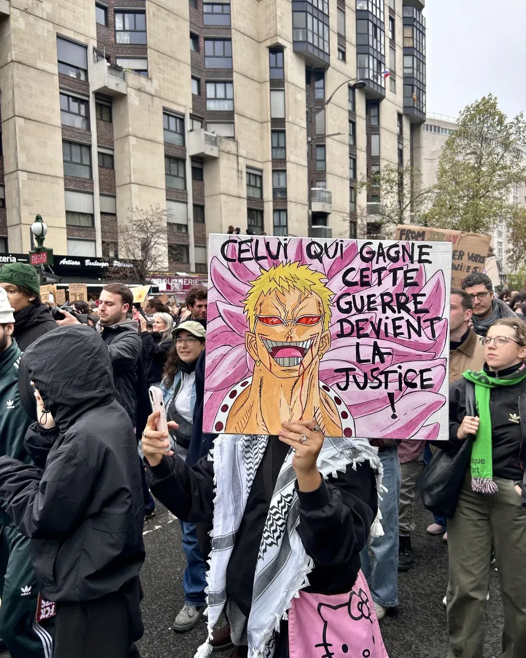 Une manifestante en keffieh, sac Hello Kitty rose à l’épaule, brandit une pancarte peinte avec Doflamingo de One Piece et le texte « Celui qui gagne cette guerre devient la justice ! »