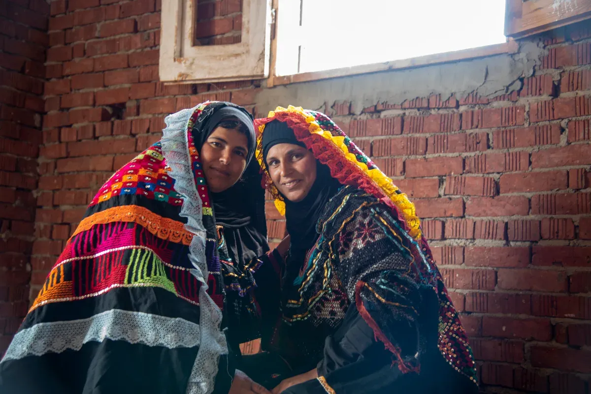Deux amies souriantes assises côte à côte, vêtues de costumes traditionnels égyptiens brodés de rouge, jaune et vert, devant un mur de briques