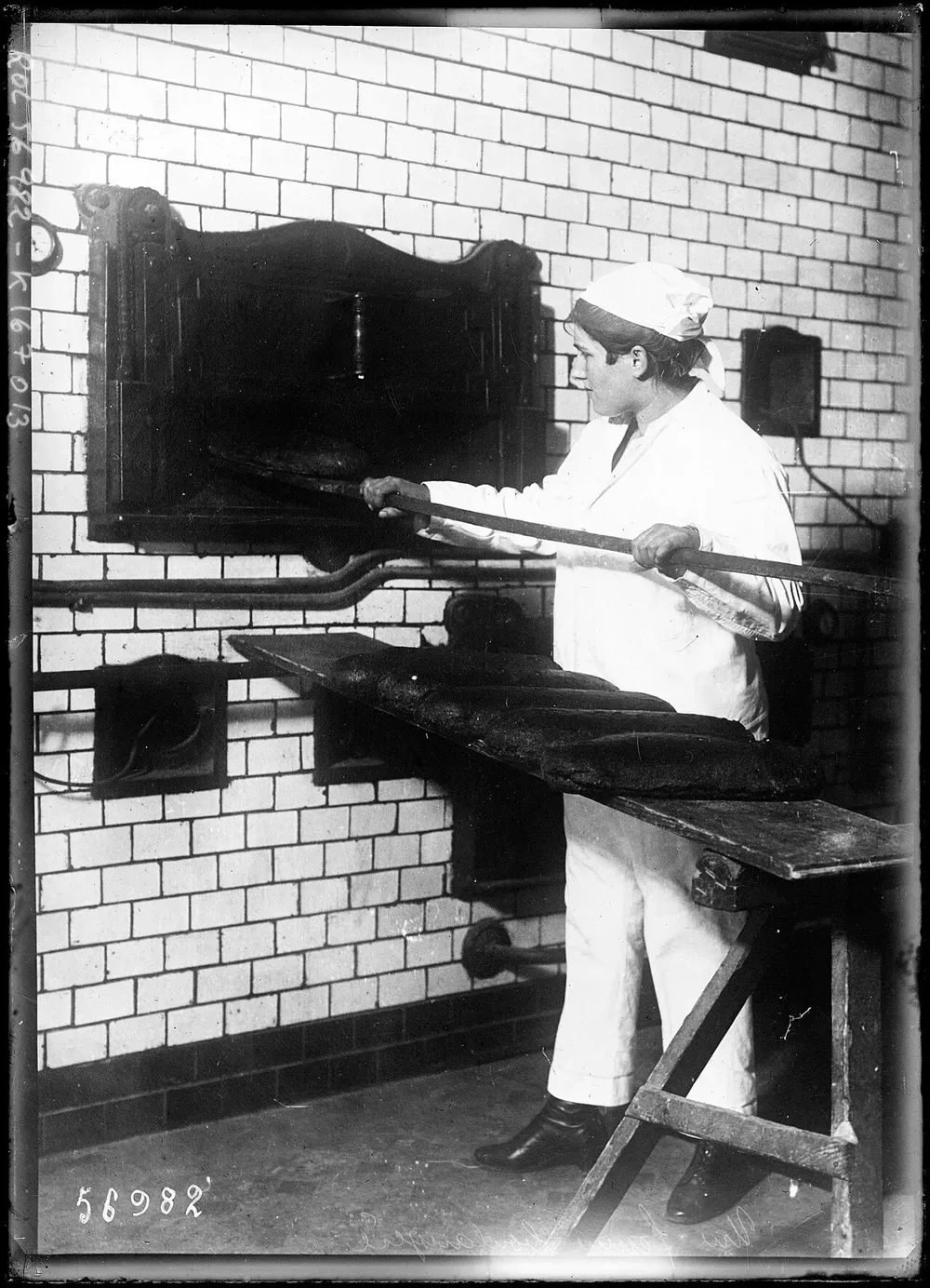 Photographie en noir et blanc d’une femme en tenue de boulangerie blanche enfournant des pains à la pelle devant un four à pain carrelé