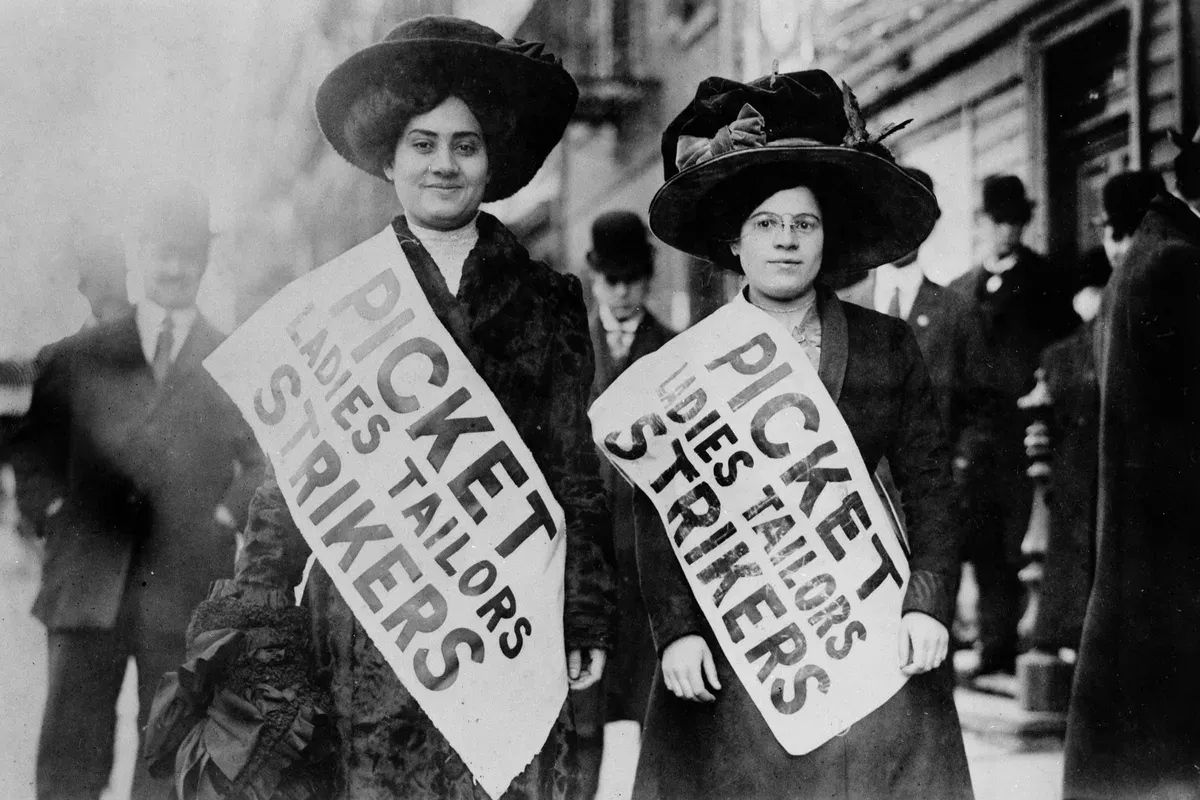 Deux grévistes new-yorkaises en manteau et grand chapeau, portant des écharpes imprimées “Picket Ladies Tailors Strikers”