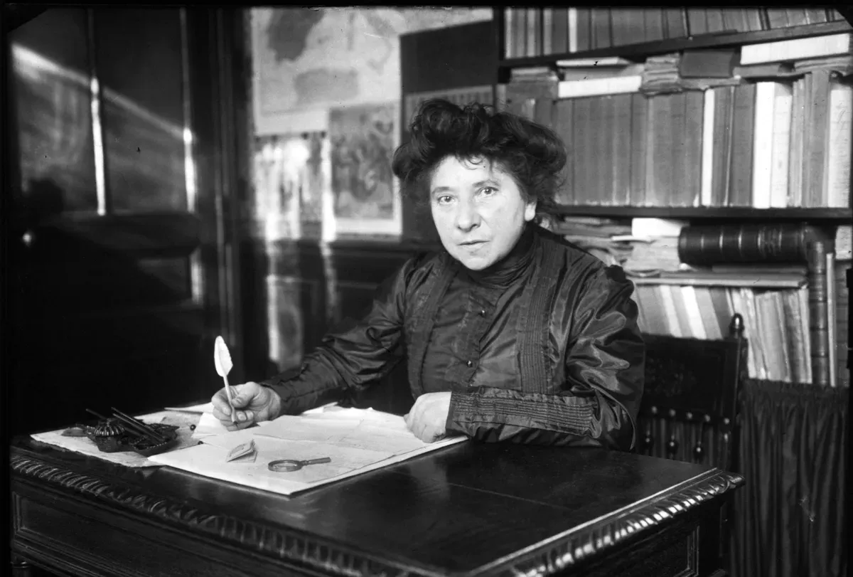 Hubertine Auclert assise à son bureau, plume à la main devant un grand registre ouvert, bibliothèque derrière elle