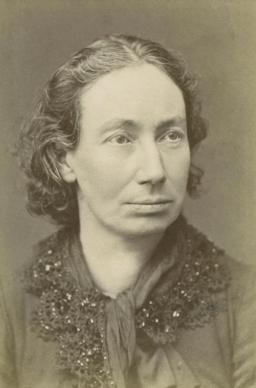 Portrait photographique de Louise Michel de face, cheveux bouclés courts, robe sombre à col brodé