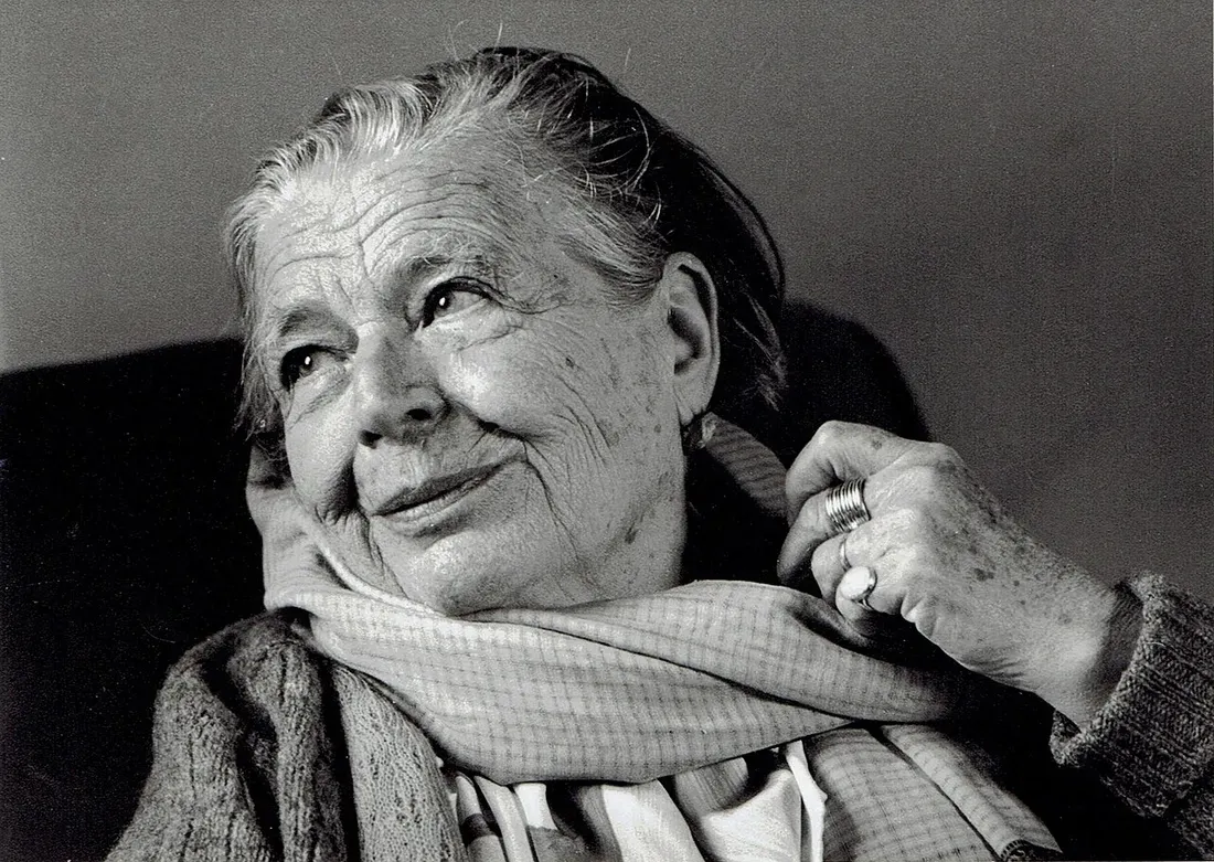 Portrait en noir et blanc de Marguerite Yourcenar âgée, souriante, le regard levé, en écharpe claire et gilet de laine