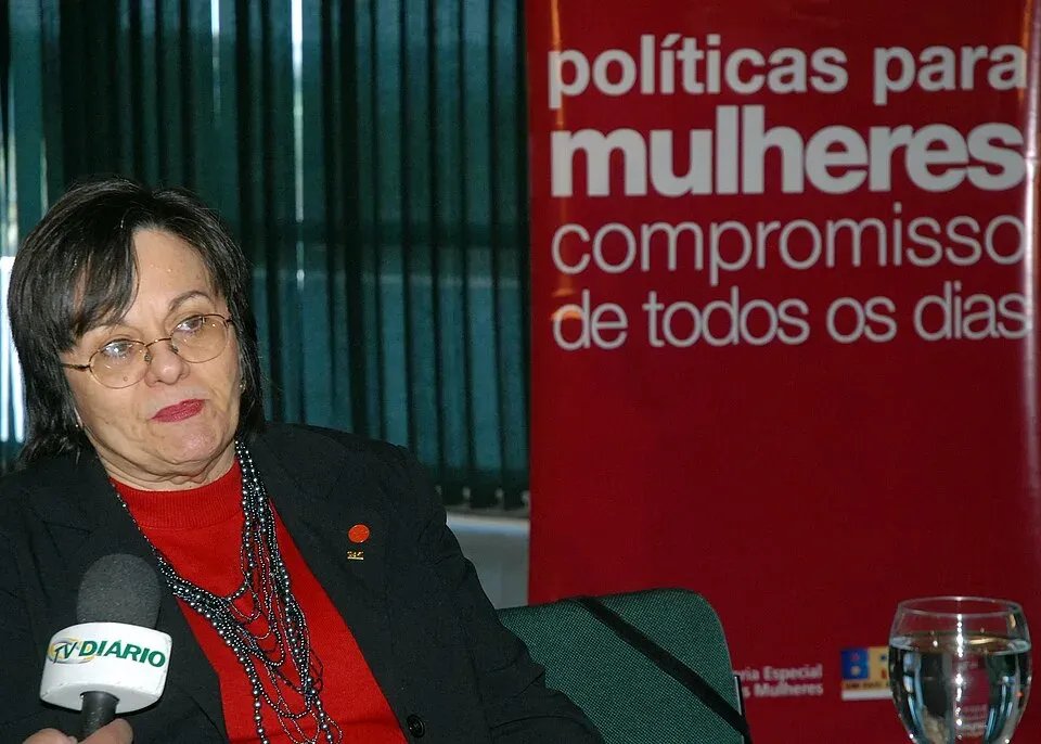 Maria da Penha, lunettes et pull rouge, répond à une interview devant une banderole « políticas para mulheres, compromisso de todos os dias »