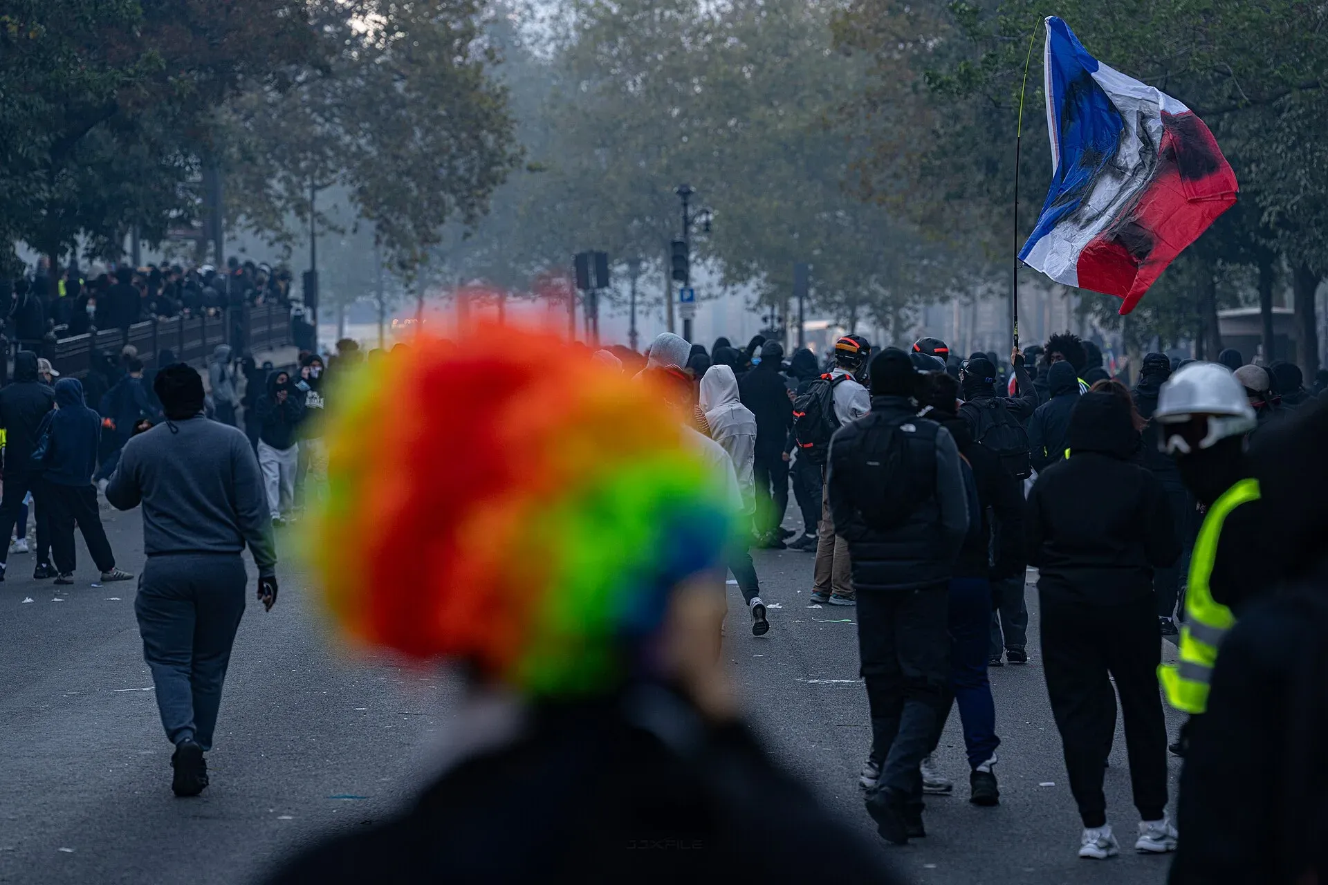 Manifestants place de la République à Paris dans la brume des gaz lacrymogènes, une perruque arc-en-ciel au premier plan et un drapeau français noirci en arrière-plan