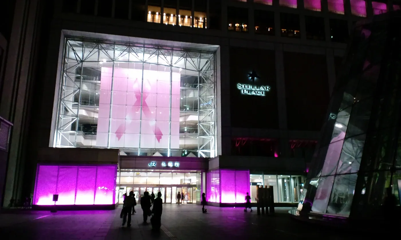 Immense ruban rose illuminé sur la façade vitrée de la gare de Sapporo la nuit, avec des passants en contrebas