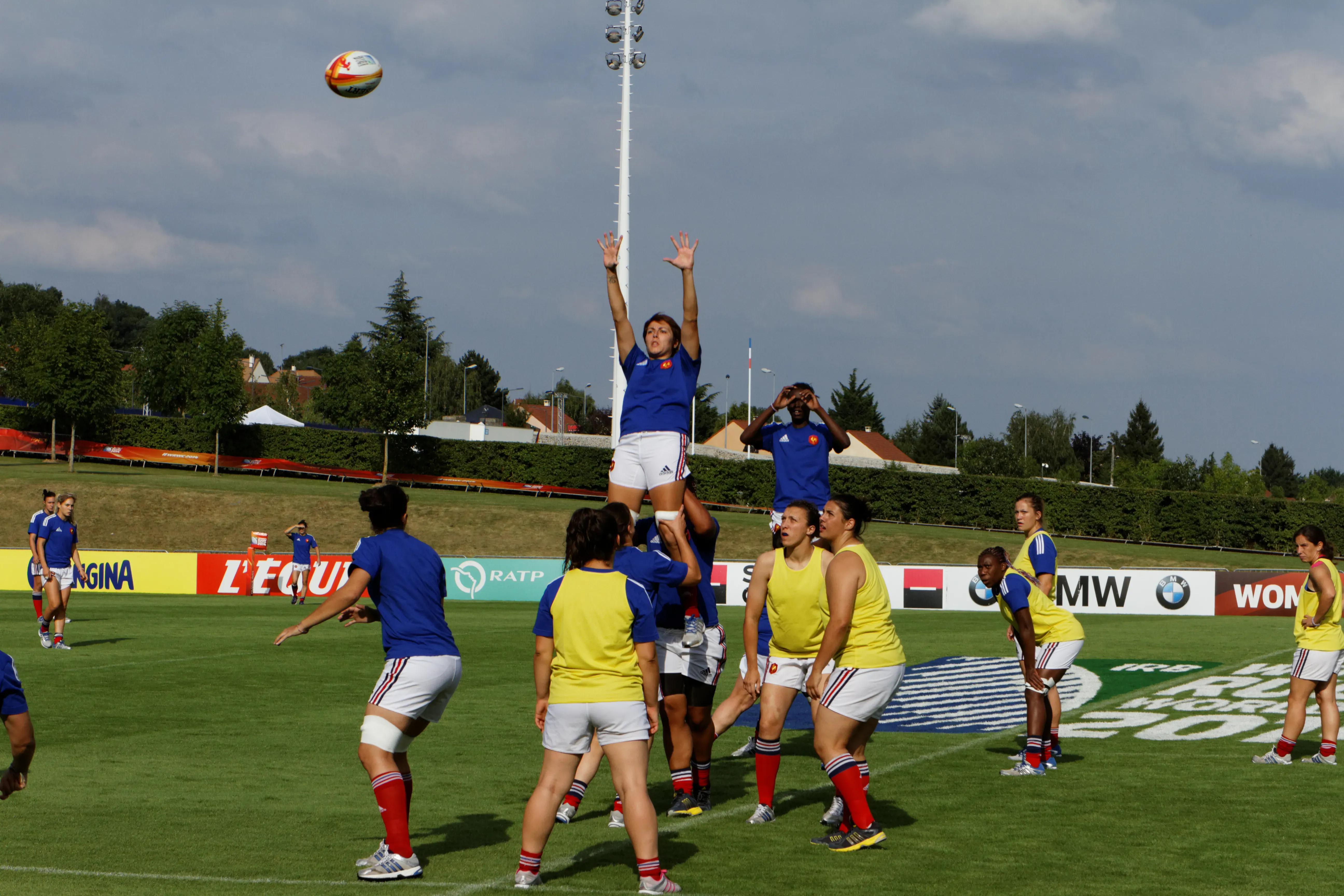 Des joueuses de l’équipe de France de rugby à XV sautent pour un touché lors d’un entraînement de la Coupe du monde 2014, une bannière du journal L’Équipe visible en arrière-plan du terrain