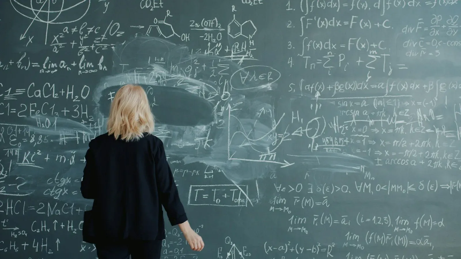 Une scientifique de dos devant un grand tableau noir couvert de formules de maths écrites à la craie