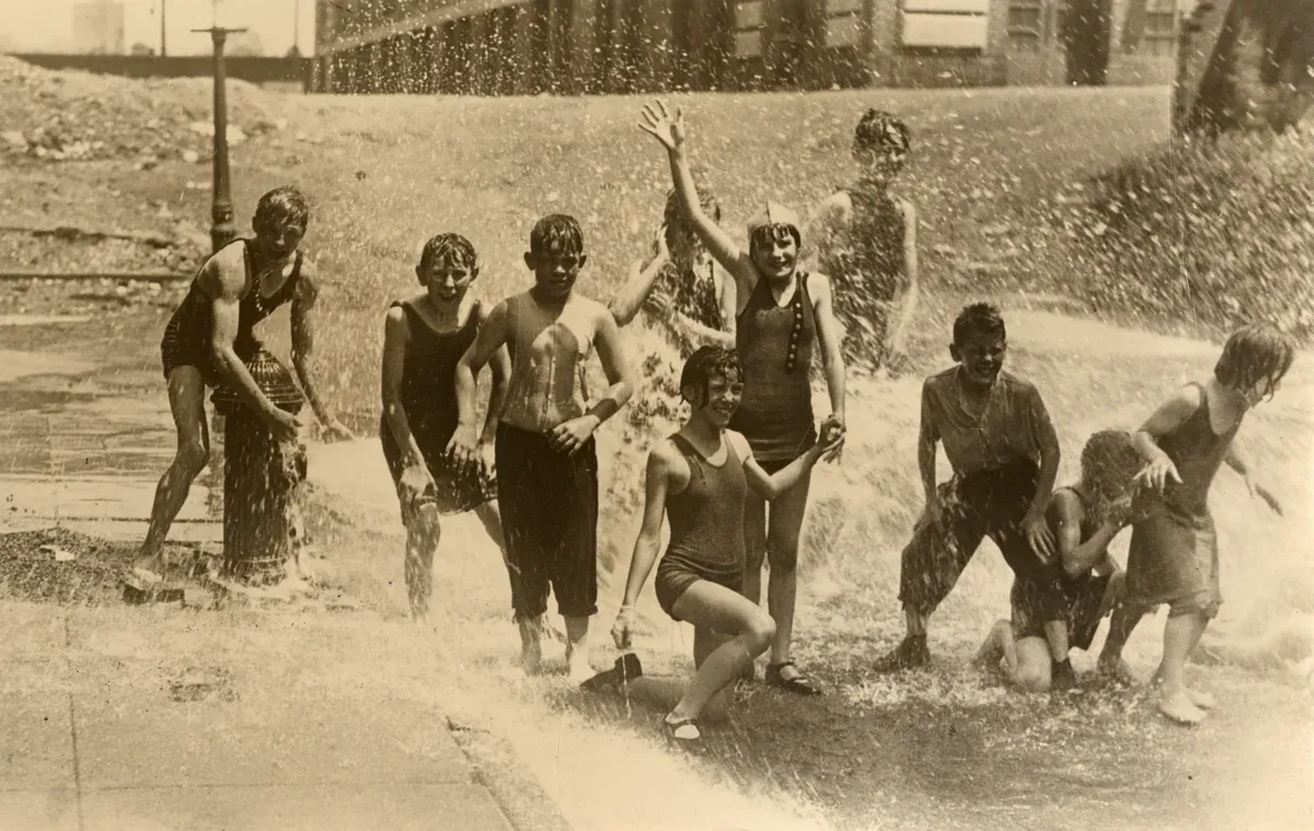 Groupe d’enfants en maillot riant sous le jet d’eau d’une bouche d’incendie dans une rue, photographie sépia des années 1920
